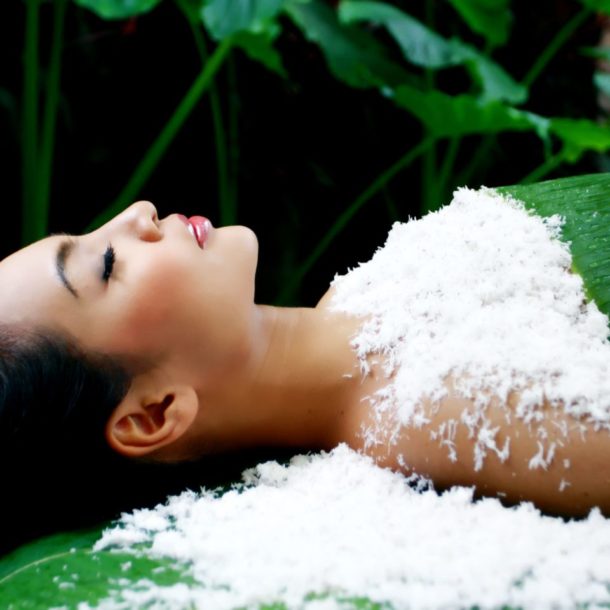 Woman covered in shaved coconut, surrounded by growing plants on green massage bed.
