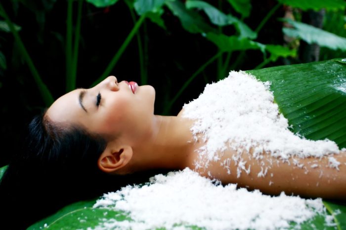 Woman covered in shaved coconut, surrounded by growing plants on green massage bed.