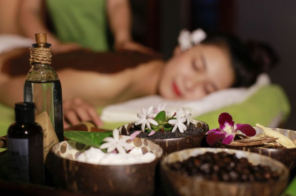Woman receiving massage in background while essential oils and aromatics are shown in foreground.